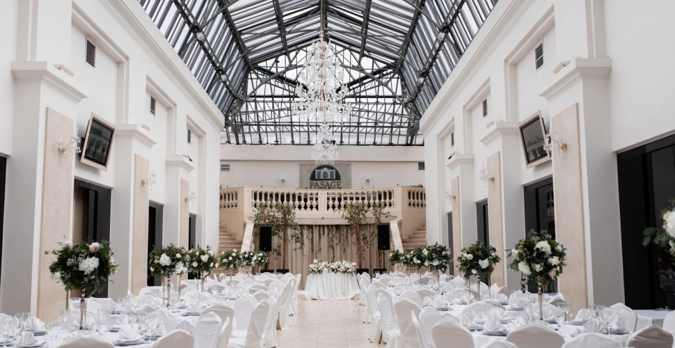 Elegant wedding reception setup inside a bright glass-roofed event space St. Louis with white decor, chandeliers, and floral centerpieces.
