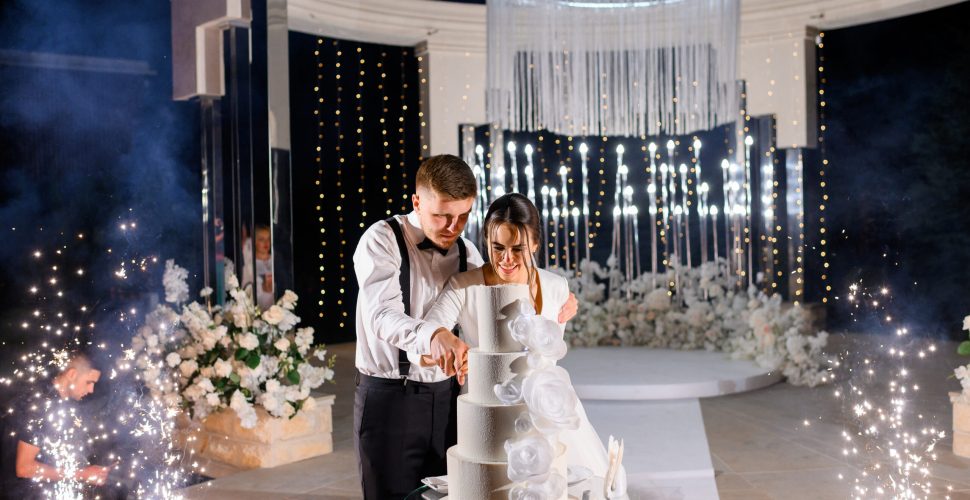 Front view happy married couple loving man and woman cutting big white cake on wedding ceremony at night. Standing near well-decorated luxury altar with flowers and sparklers. Special event